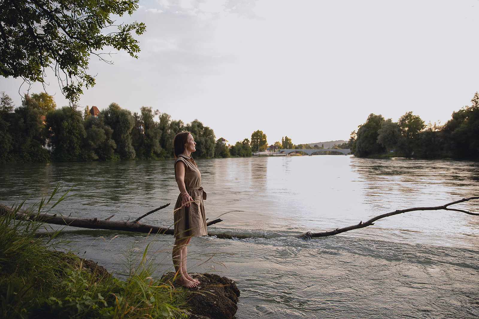 Elvira Christina Wettstein steht mit ausgebreiteten Armen am Ufer eines Flusses.
